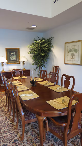 Long wooden dining table with chairs, placemats, lamps, framed art, and a plant.