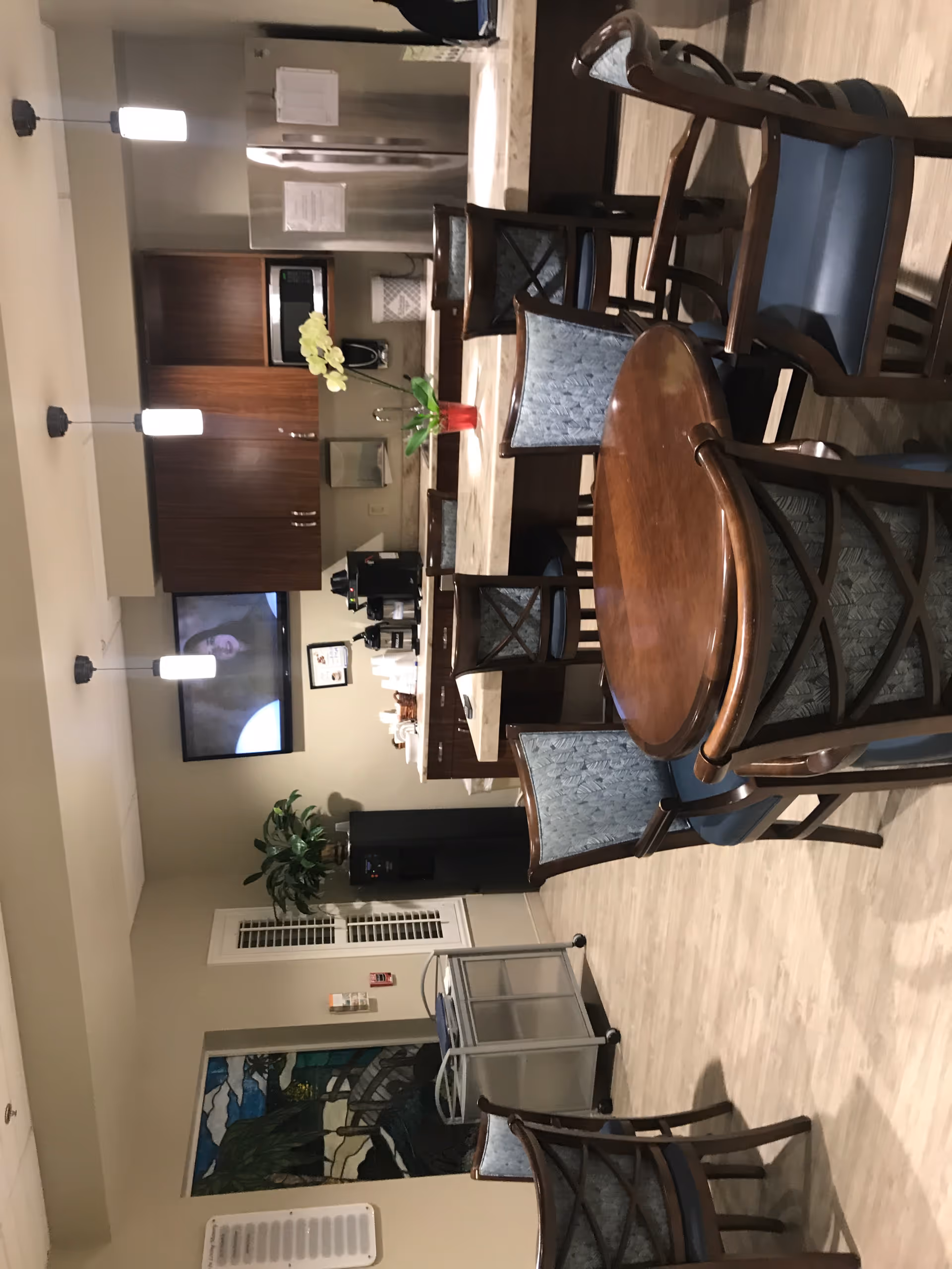 Interior communal dining area with round wooden table, blue chairs, plants, and a kitchenette.