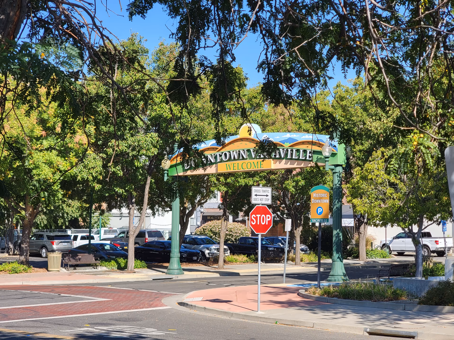 Arched welcome sign over a street with trees, benches, and parked cars.