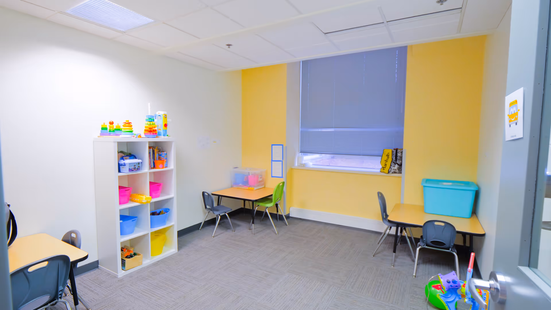 Bright children's activity room with shelves of toys, small tables, and colorful chairs.