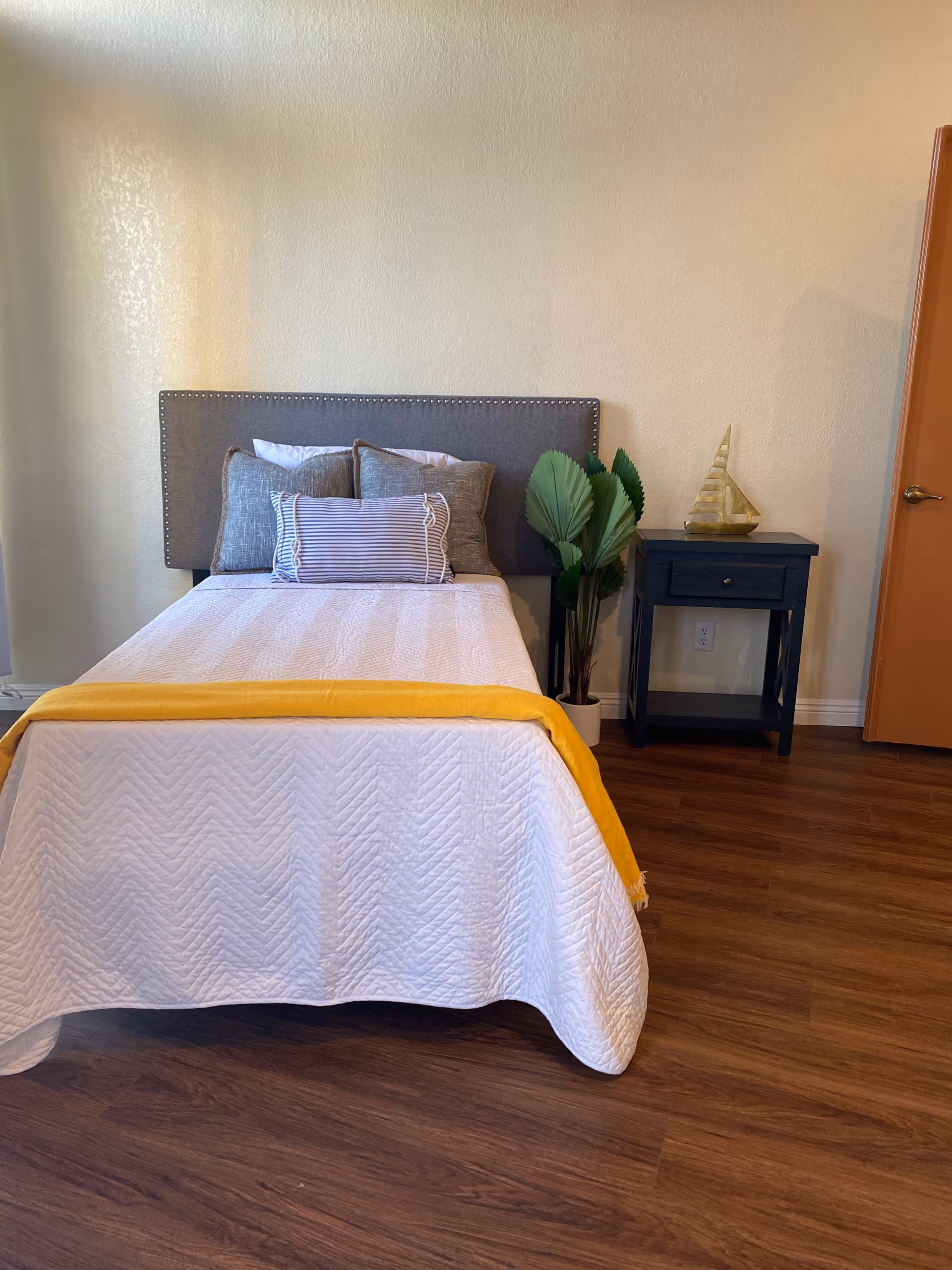 A neatly made bedroom featuring a bed, nightstand, plant, and wooden floor.