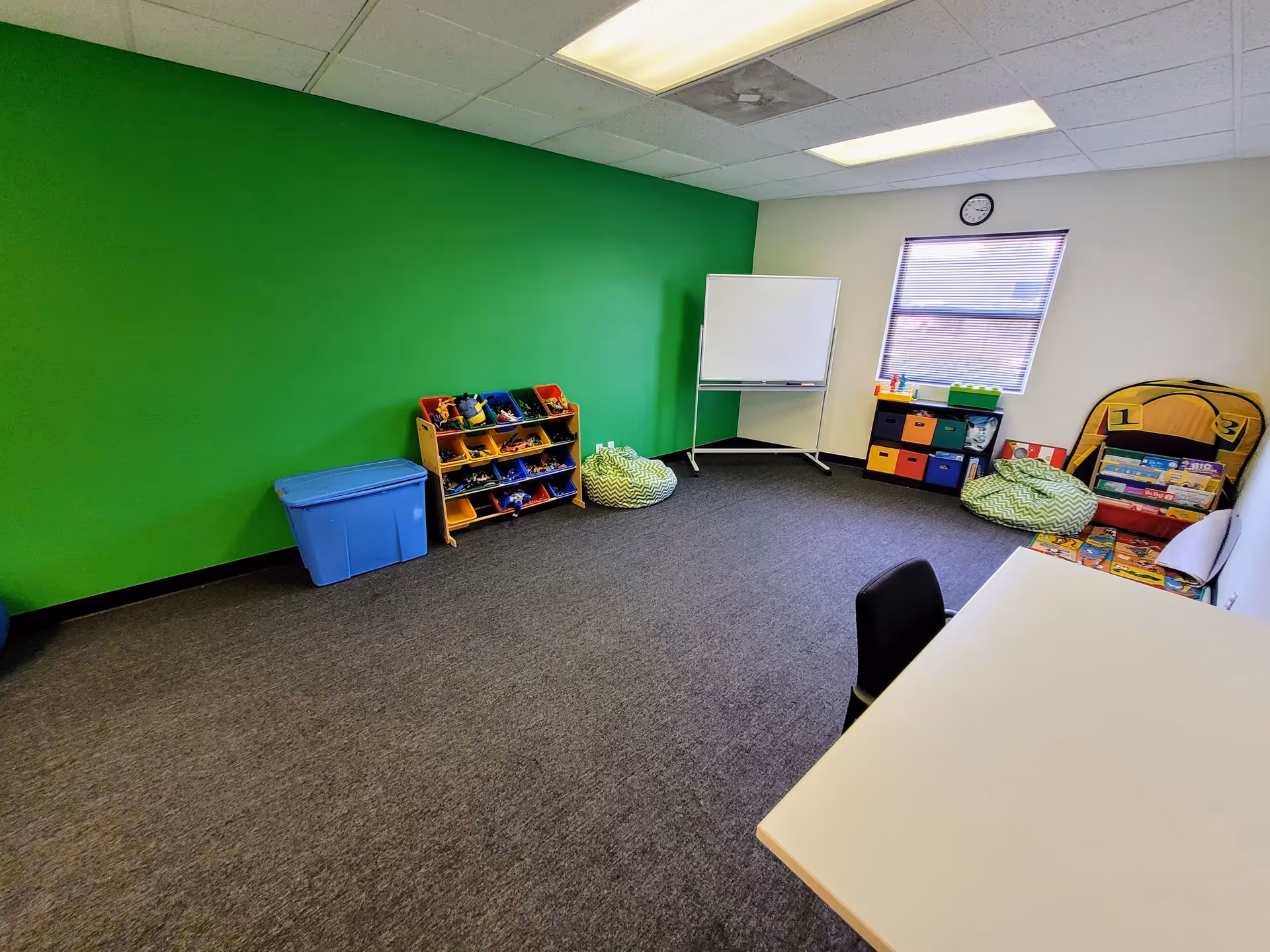 Bright activity room with green wall, toy shelves, whiteboard, bean bags, and a table.