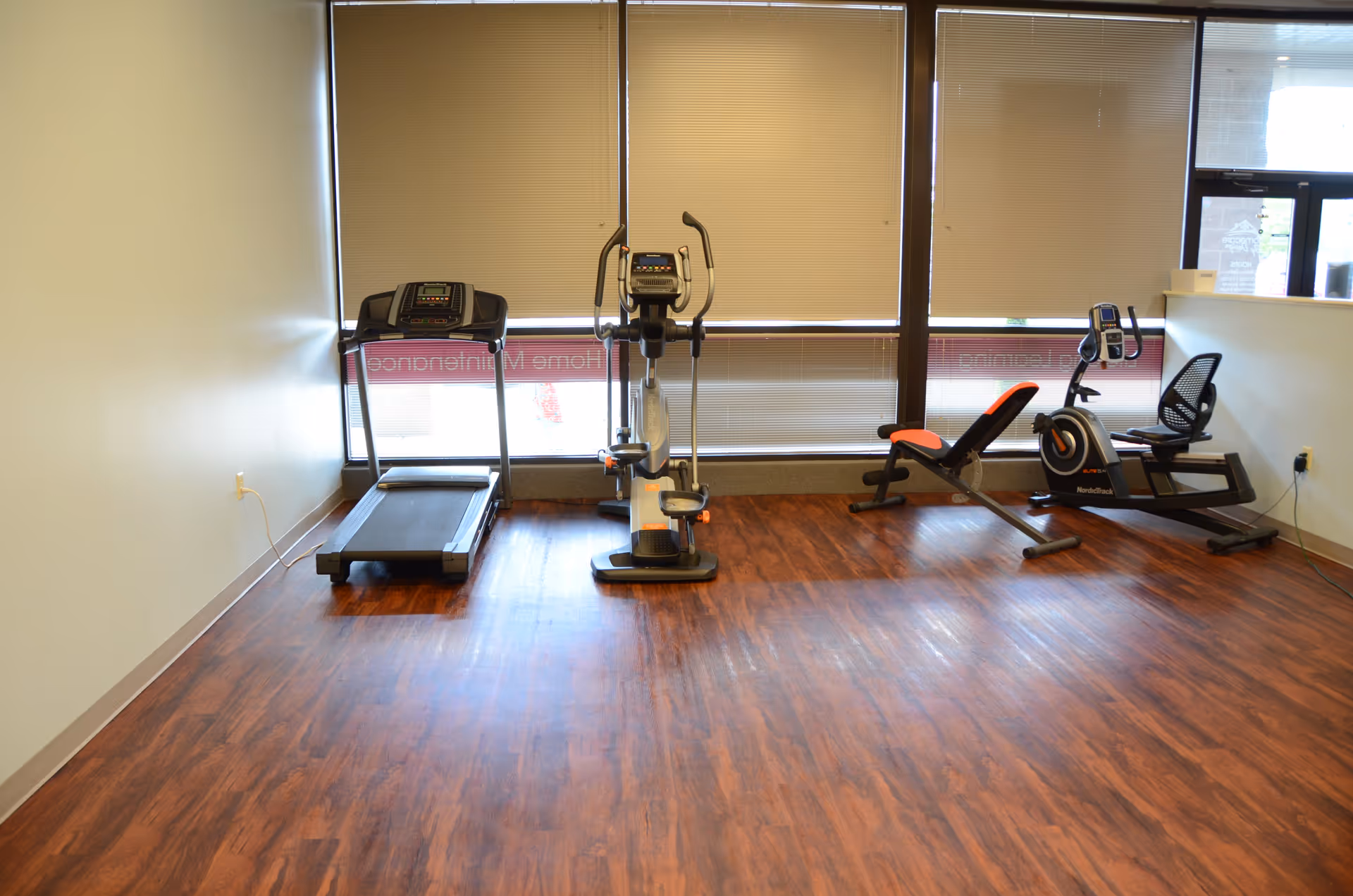Small indoor gym with treadmill, elliptical, and recumbent bike by large windows.