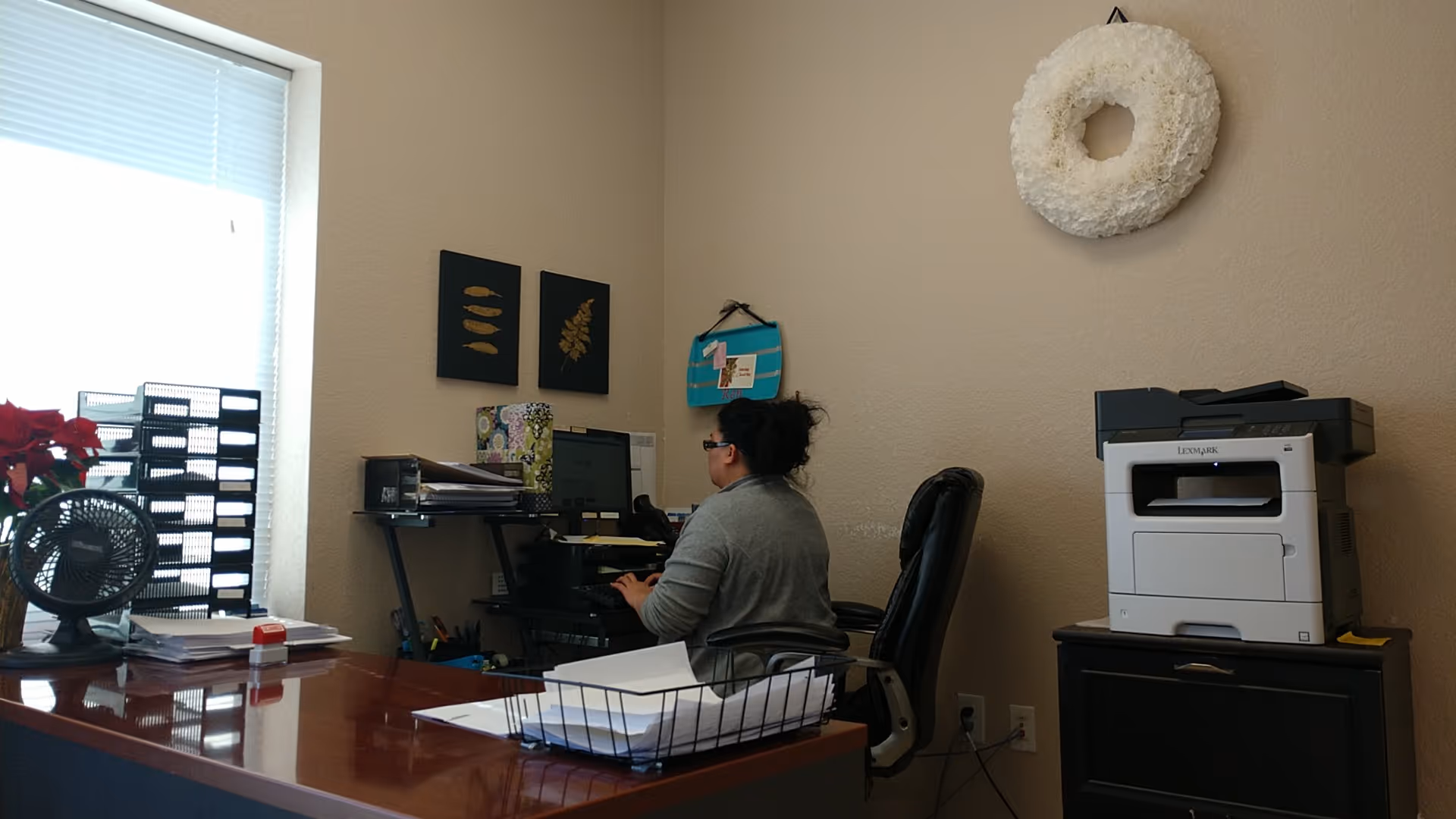 A person sits at a desk in a small office with computer and printer.