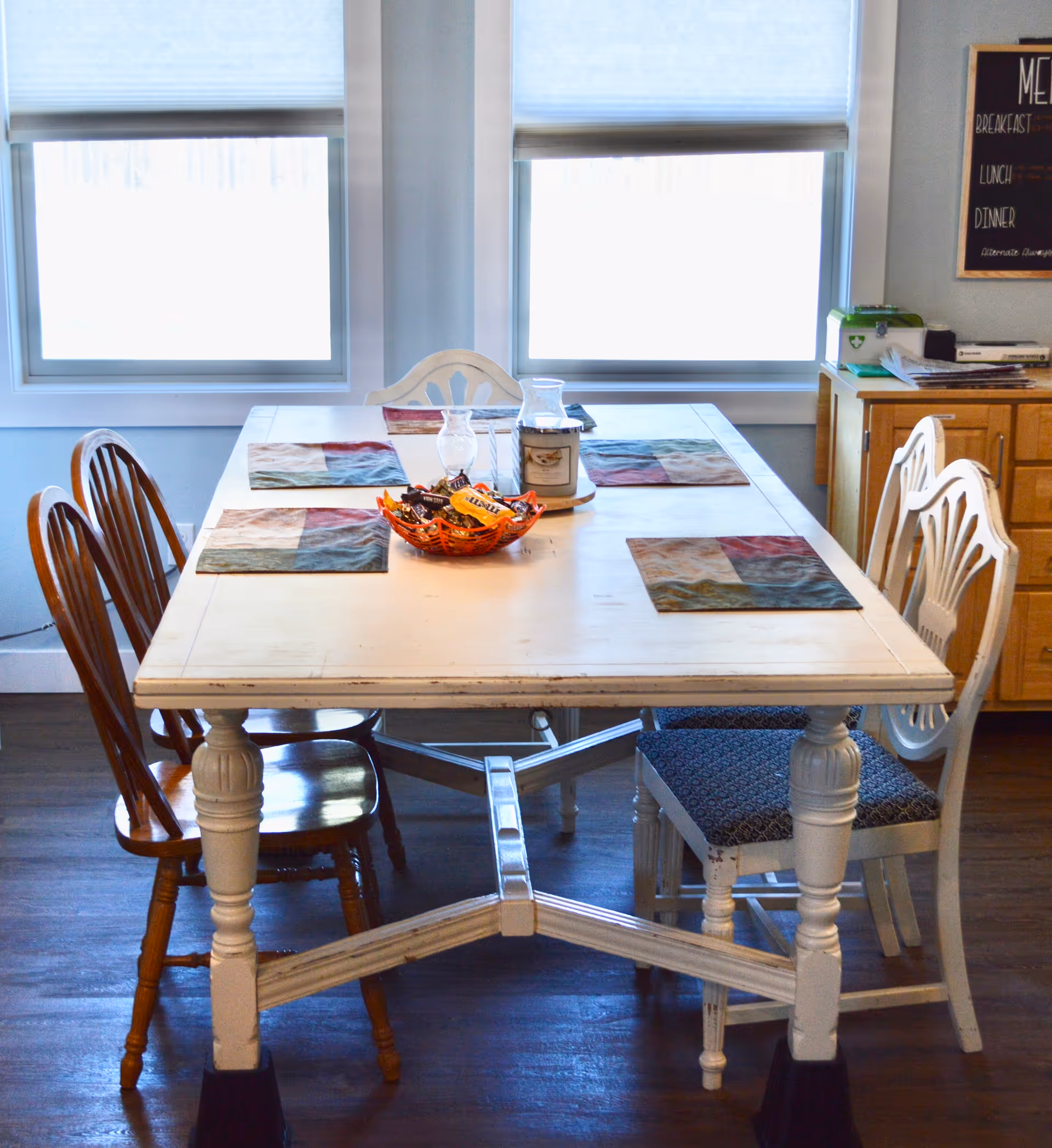 White wooden dining table with mixed chairs in a bright, window-lit dining area.