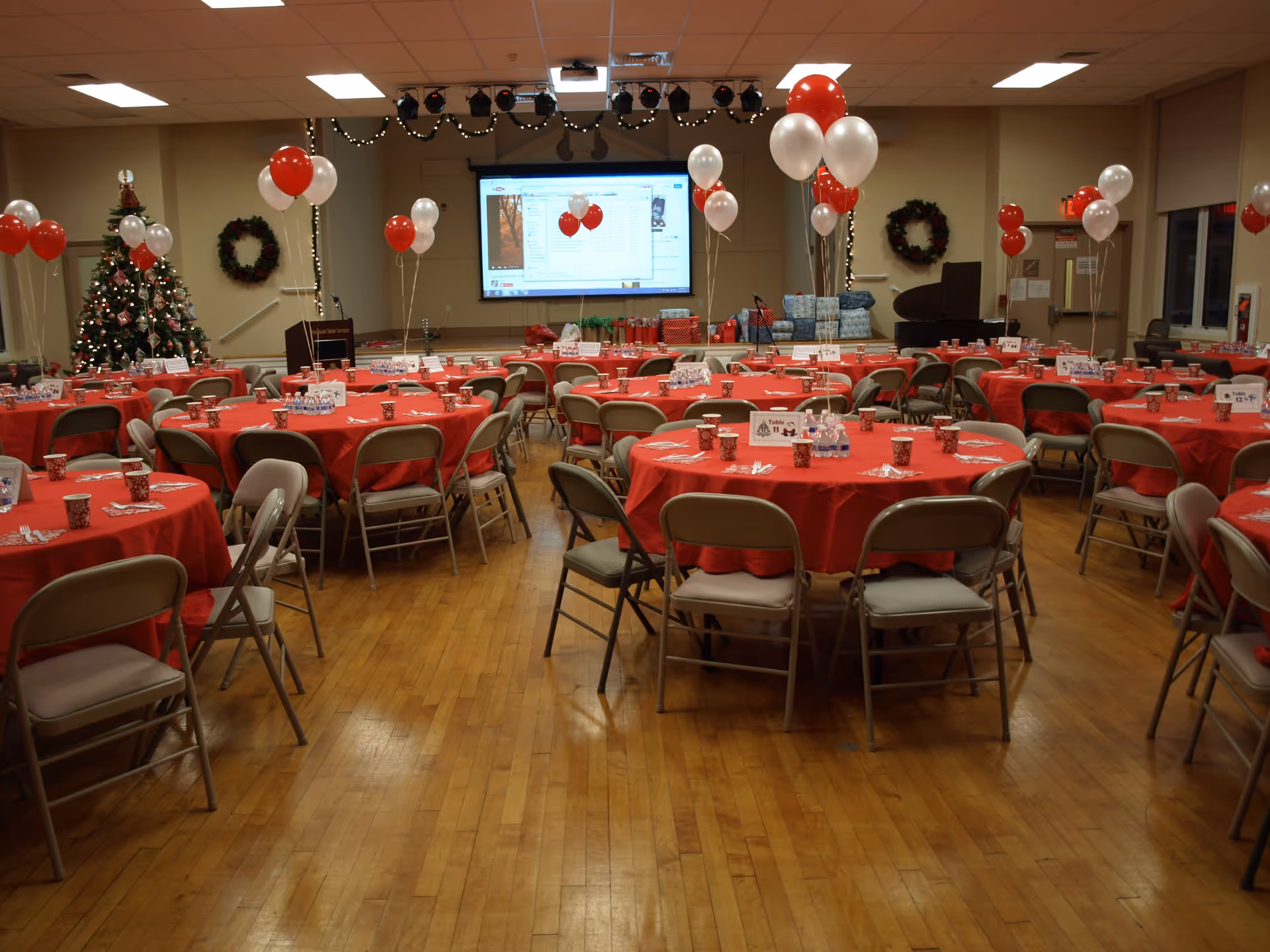 Banquet hall with red tablecloths, balloons, Christmas tree, and gifts in a decorated event space.