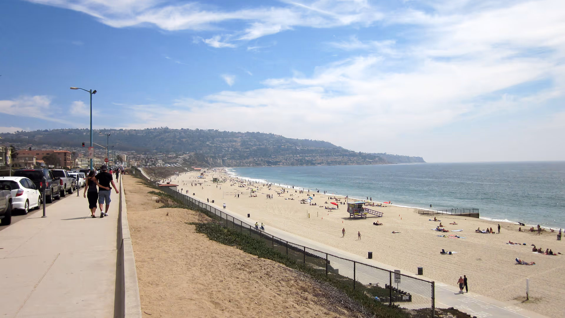 Sunlit beachfront promenade with pedestrians, cars, and expansive ocean view.