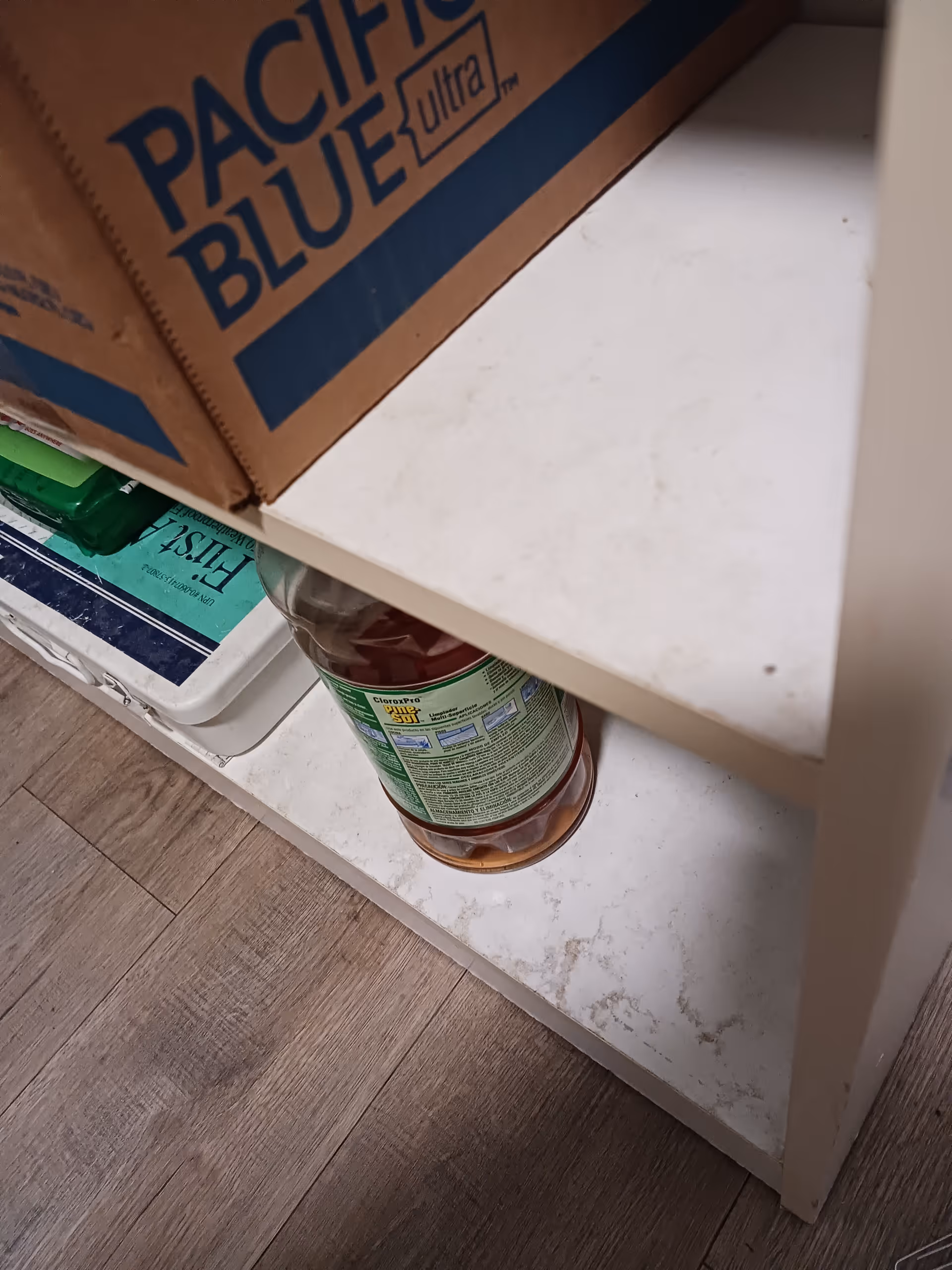 A cluttered cabinet shelf holds cleaning supplies and a boxed item labeled Pacific Blue Ultra.