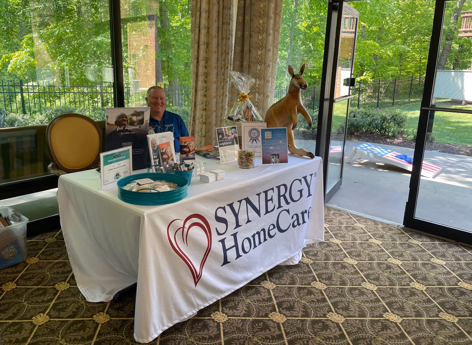 Indoors, a Synergy HomeCare display table with brochures and a smiling staff member.