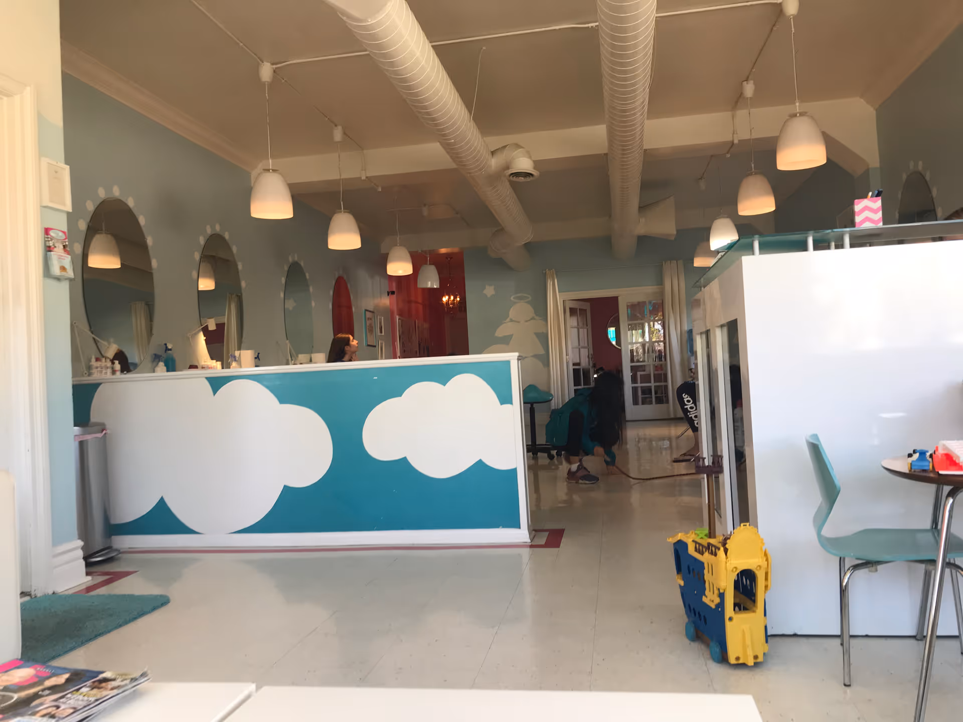 Pastel hair salon interior with cloud mural, round mirrors, reception desk, and staff at work.