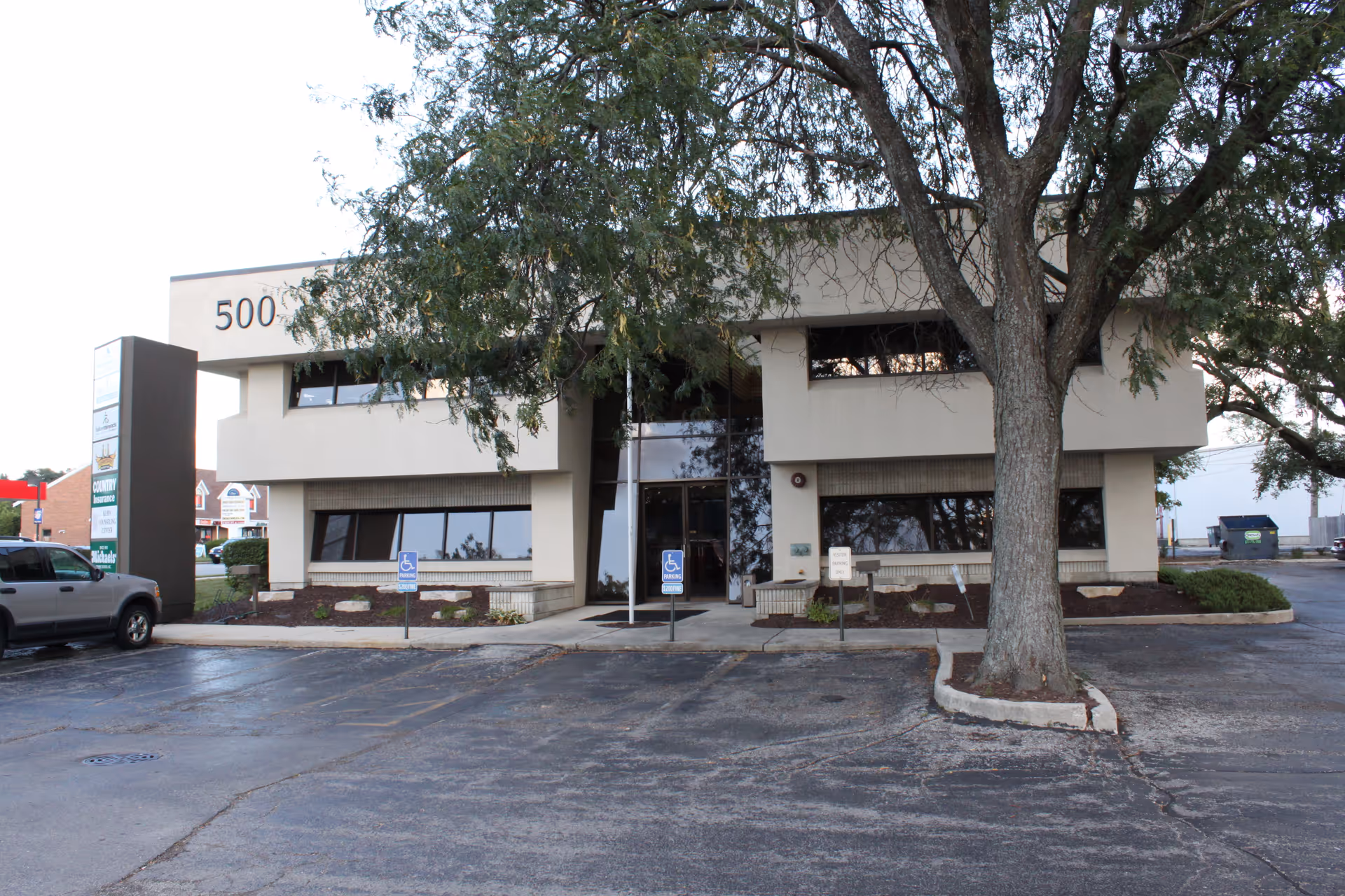Exterior of a beige two story building with a tree, parking lot, and handicap signs.