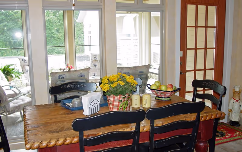 Wood dining table with chairs, flowers, fruit basket, and large windows in bright dining area.