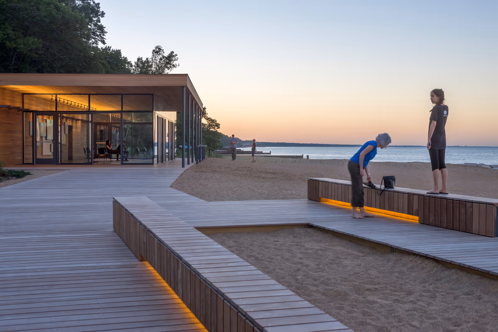Glass-walled facility with wooden boardwalk along a sandy beach at sunset.