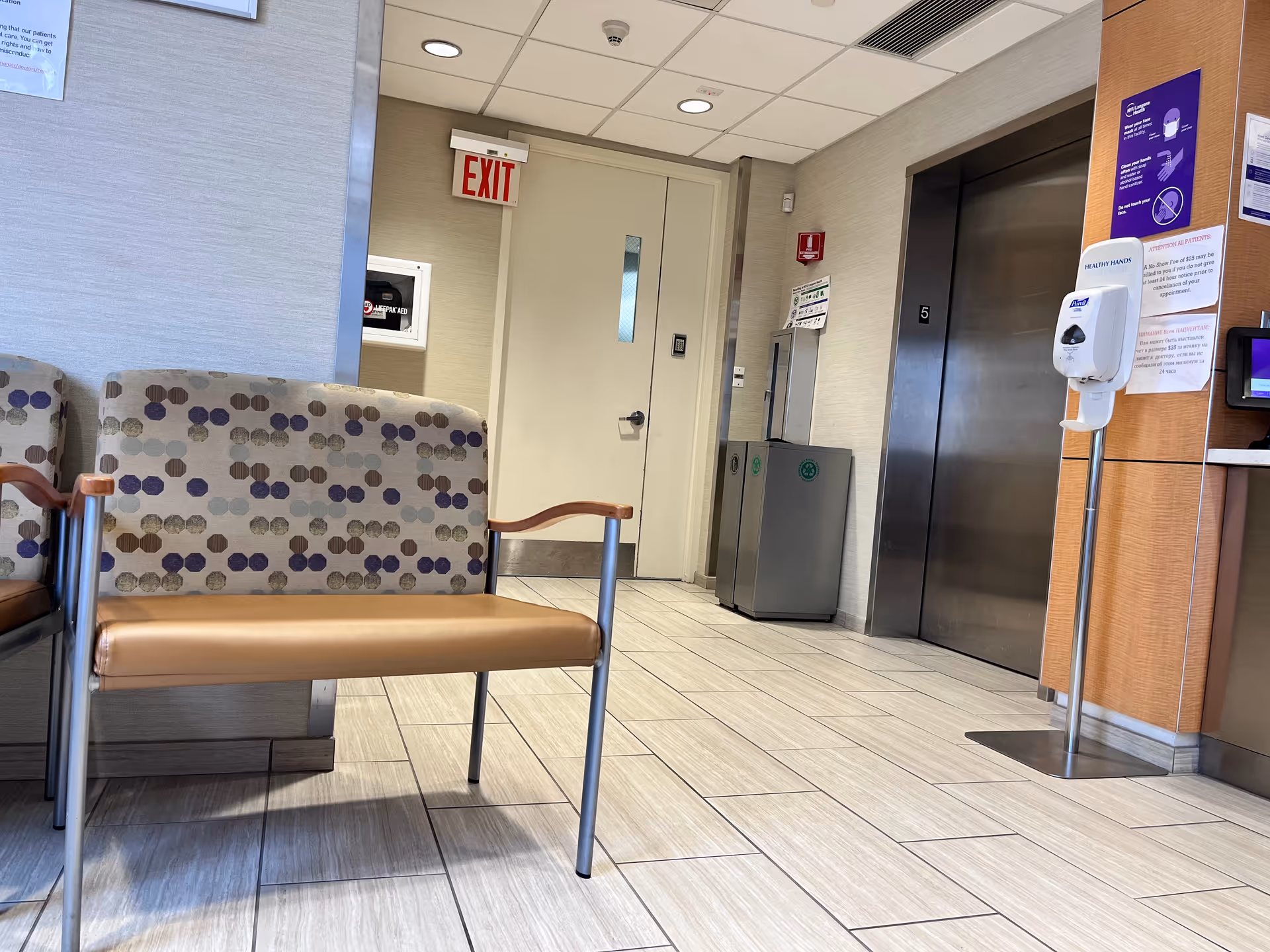 Indoor hospital-like lobby corridor with patterned chairs, elevator, exit sign, and hand sanitizer.