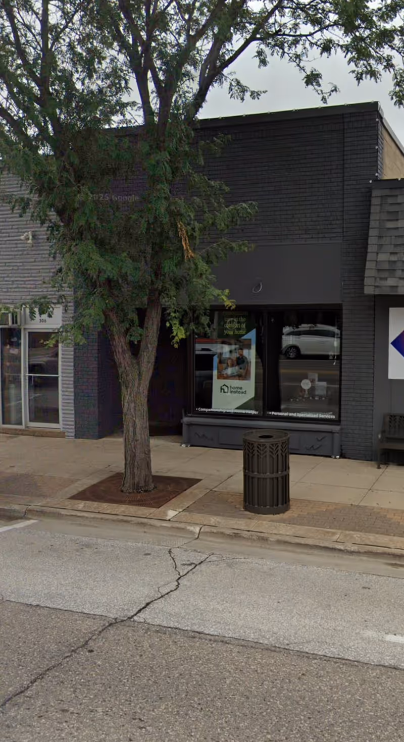 A Home Instead storefront on a tree-lined sidewalk with a poster in the window.