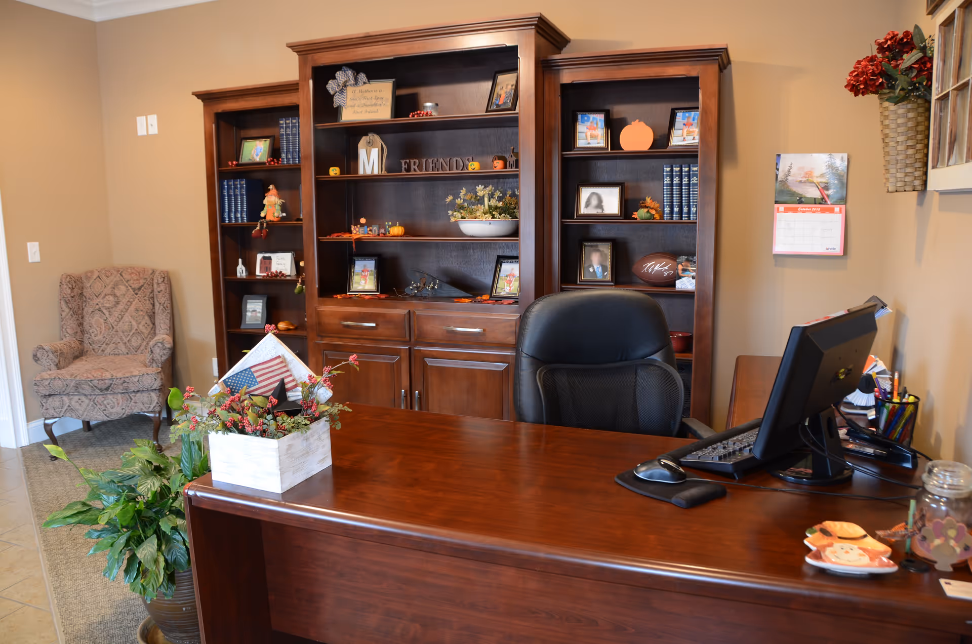 Wood reception desk with computer, bookshelves, and plants in a warm office area.