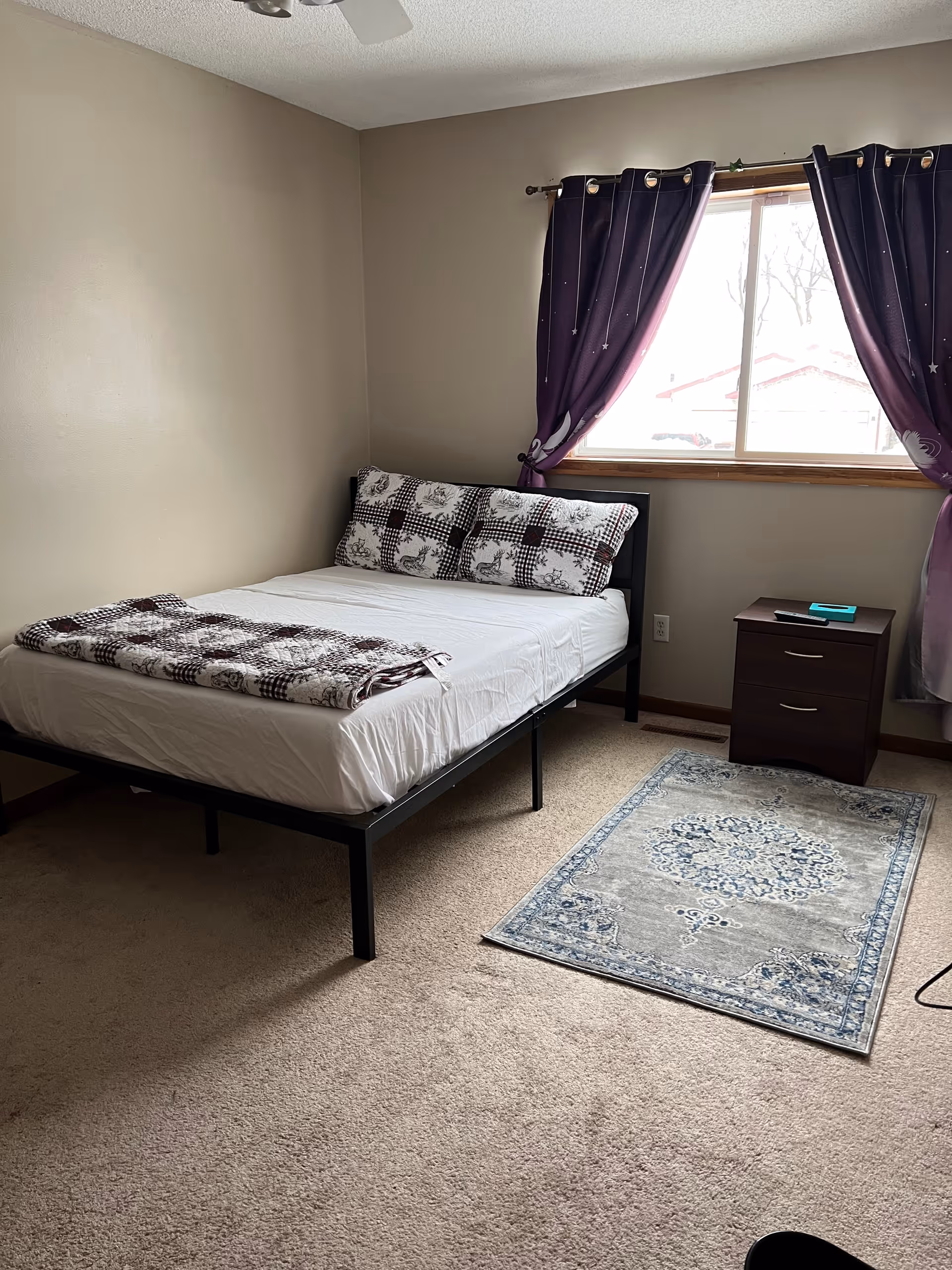 A furnished bedroom in a senior facility featuring a bed, nightstand, window with purple curtains, rug.