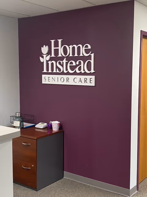 Purple wall features the Home Instead Senior Care logo with a small cabinet nearby.