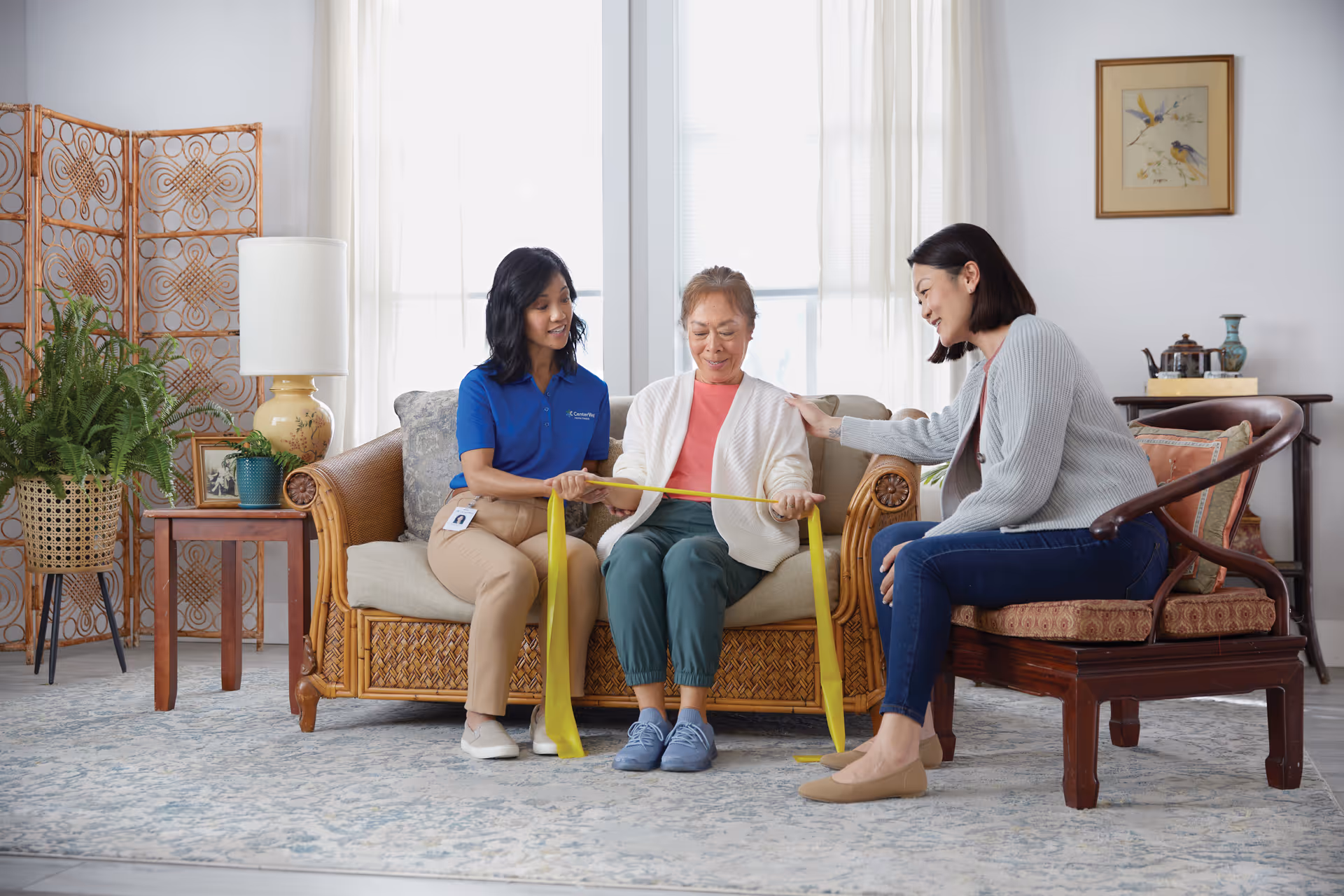 Three people sit on a living room couch, performing a seated stretch with a yellow band.