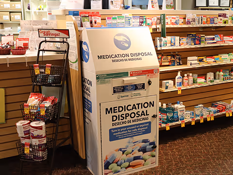 Pharmacy aisle with a large medication disposal bin and shelves of medicines.