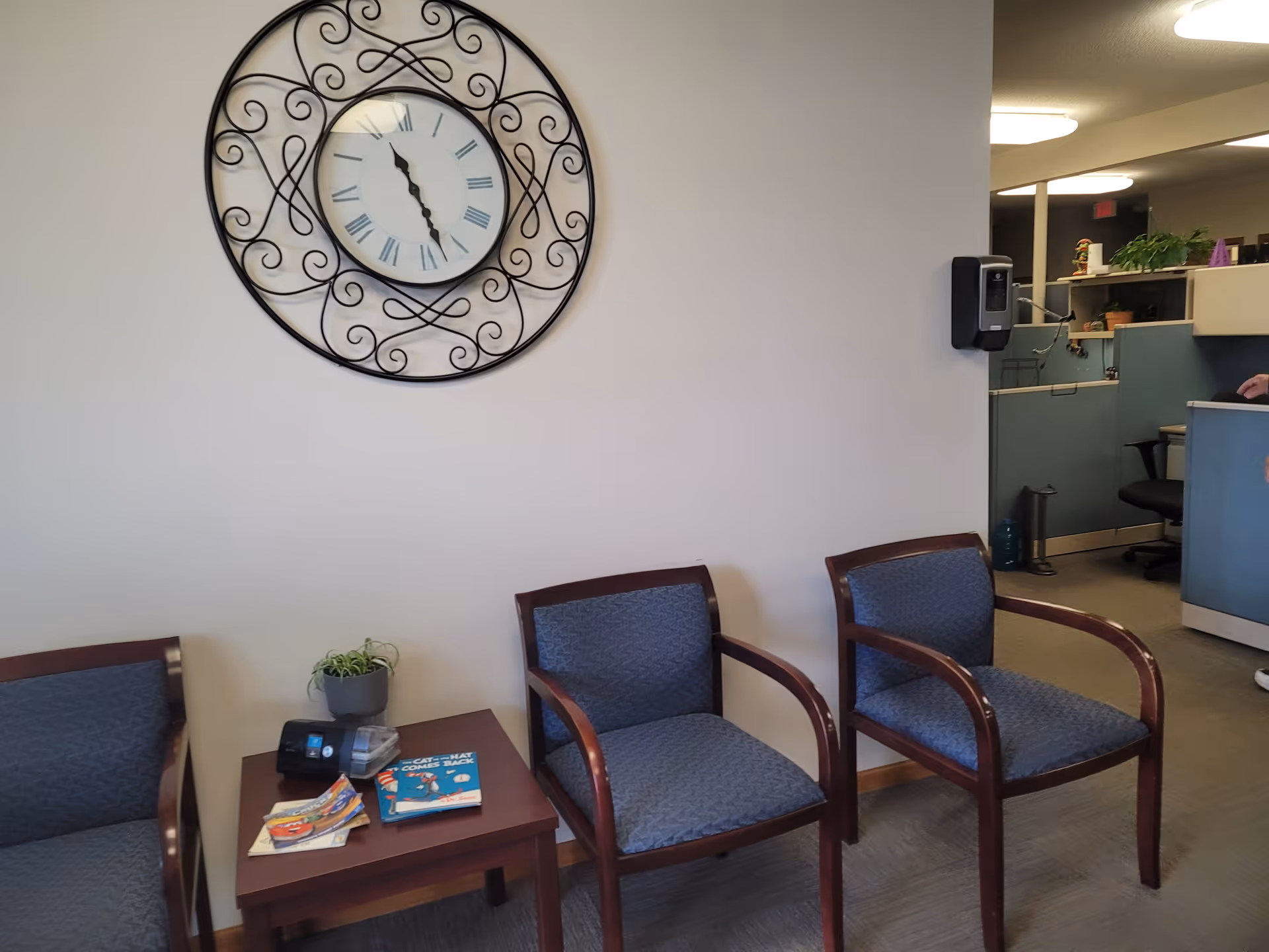 Waiting area with blue upholstered chairs, a large decorative clock, and magazines on a table.