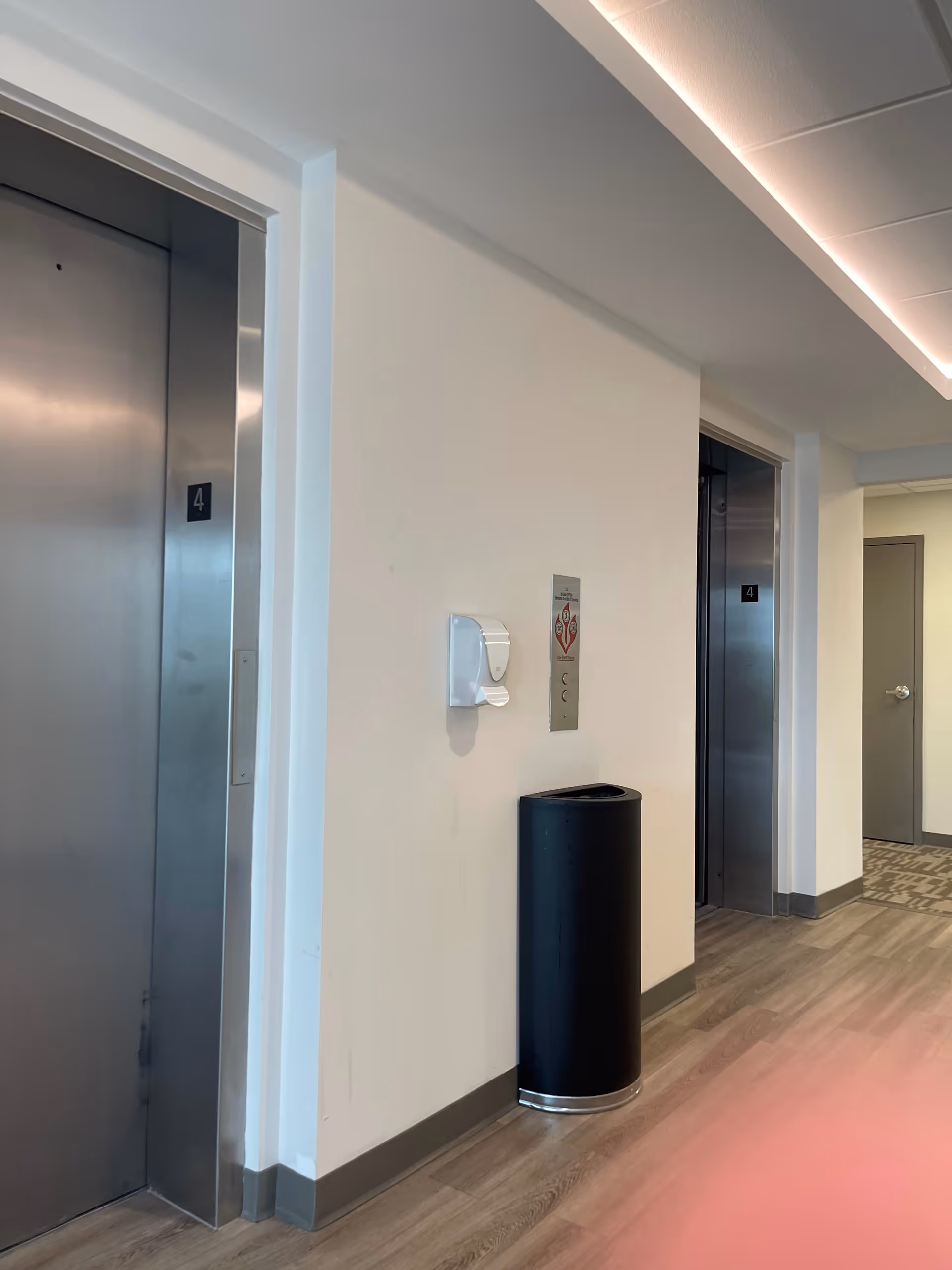Hallway interior with two elevators, a trash bin, and wall fixtures in a senior living facility.