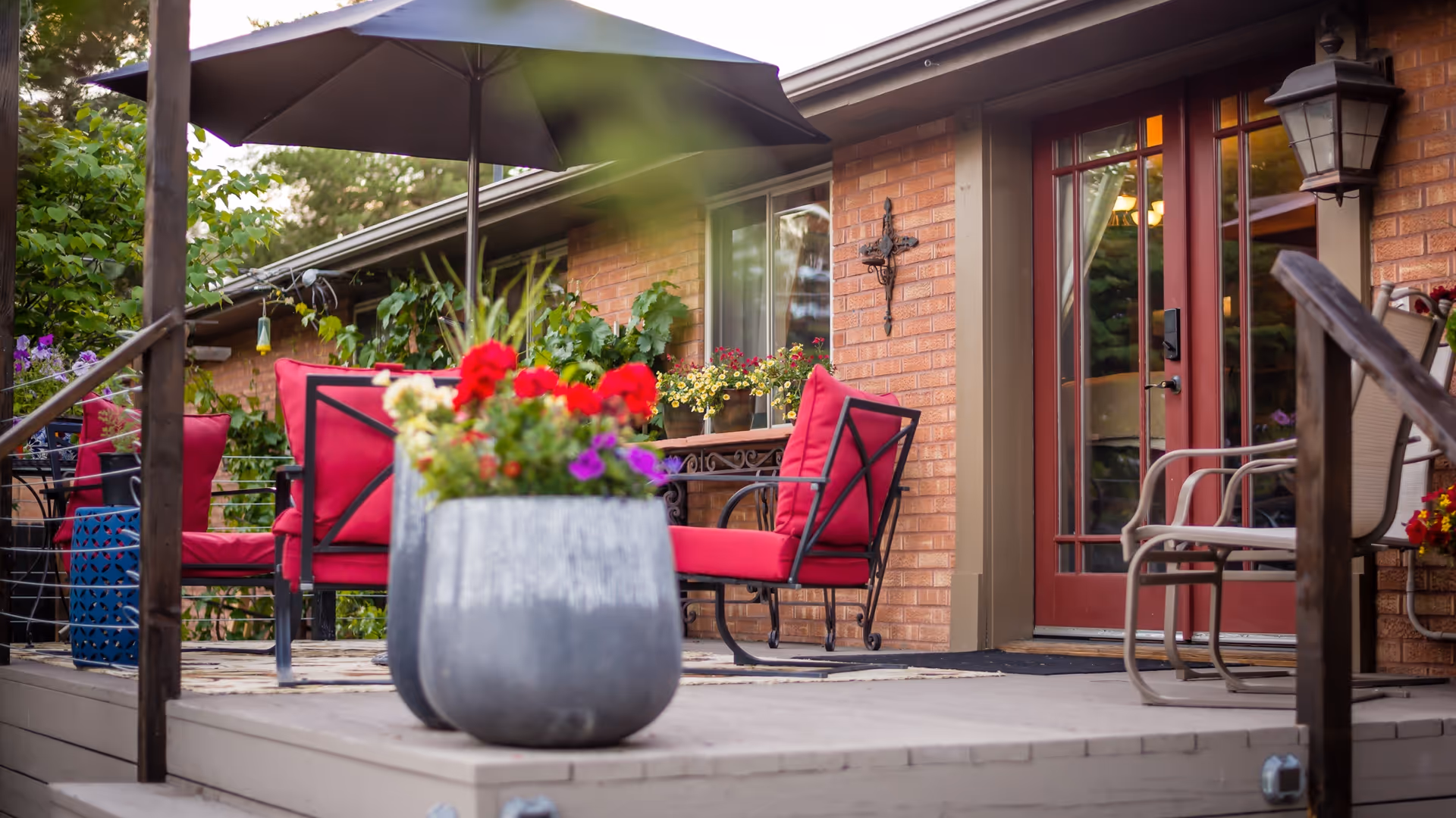 Outdoor patio with red cushioned chairs, umbrella, and potted flowers beside a brick building.