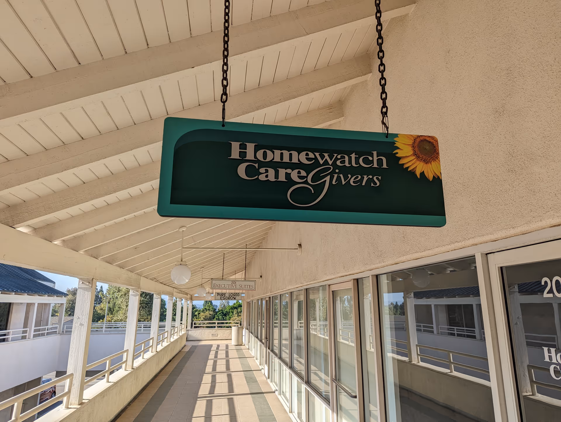 Hanging sign for Homewatch CareGivers on an exterior walkway beside glass doors.