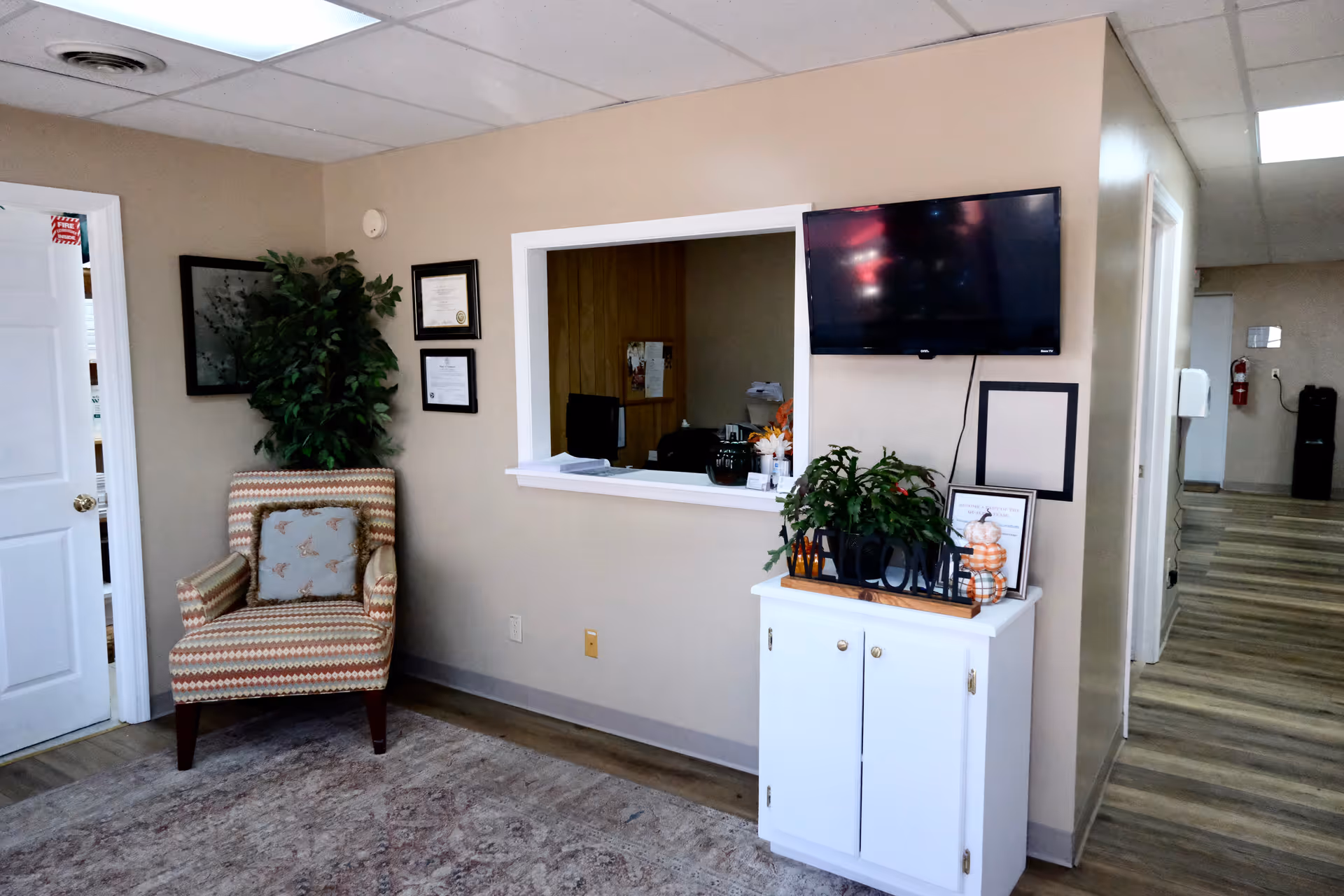 Reception lobby area with chair, plants, mounted TV, and a pass-through window.