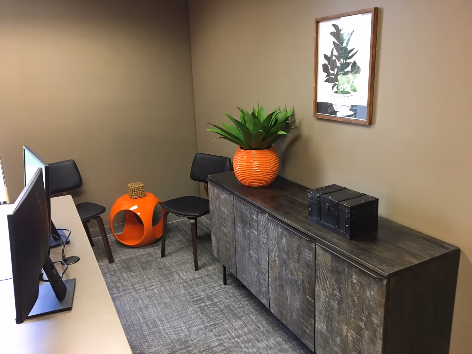 Office-style lounge with computers, a dark cabinet, orange plant pot, and wall art.