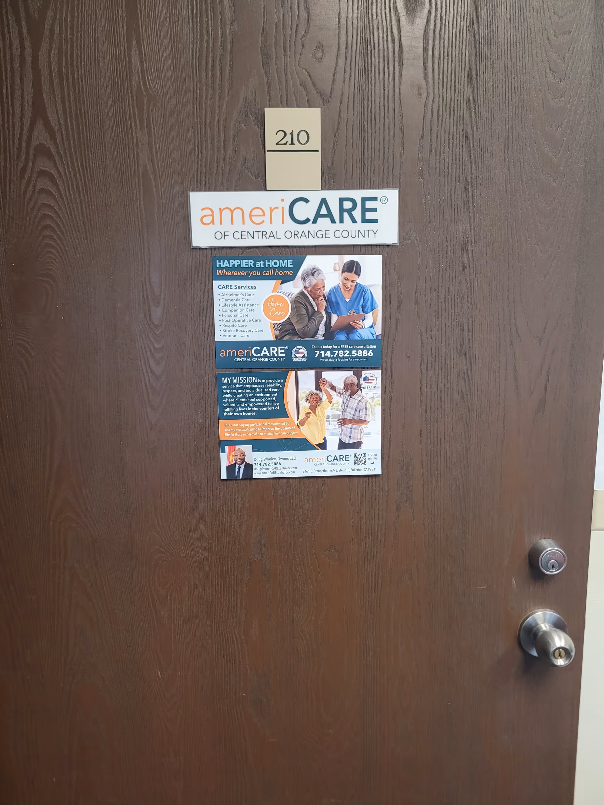 Brown wooden interior door with AmeriCARE sign and a promotional poster, room 210.