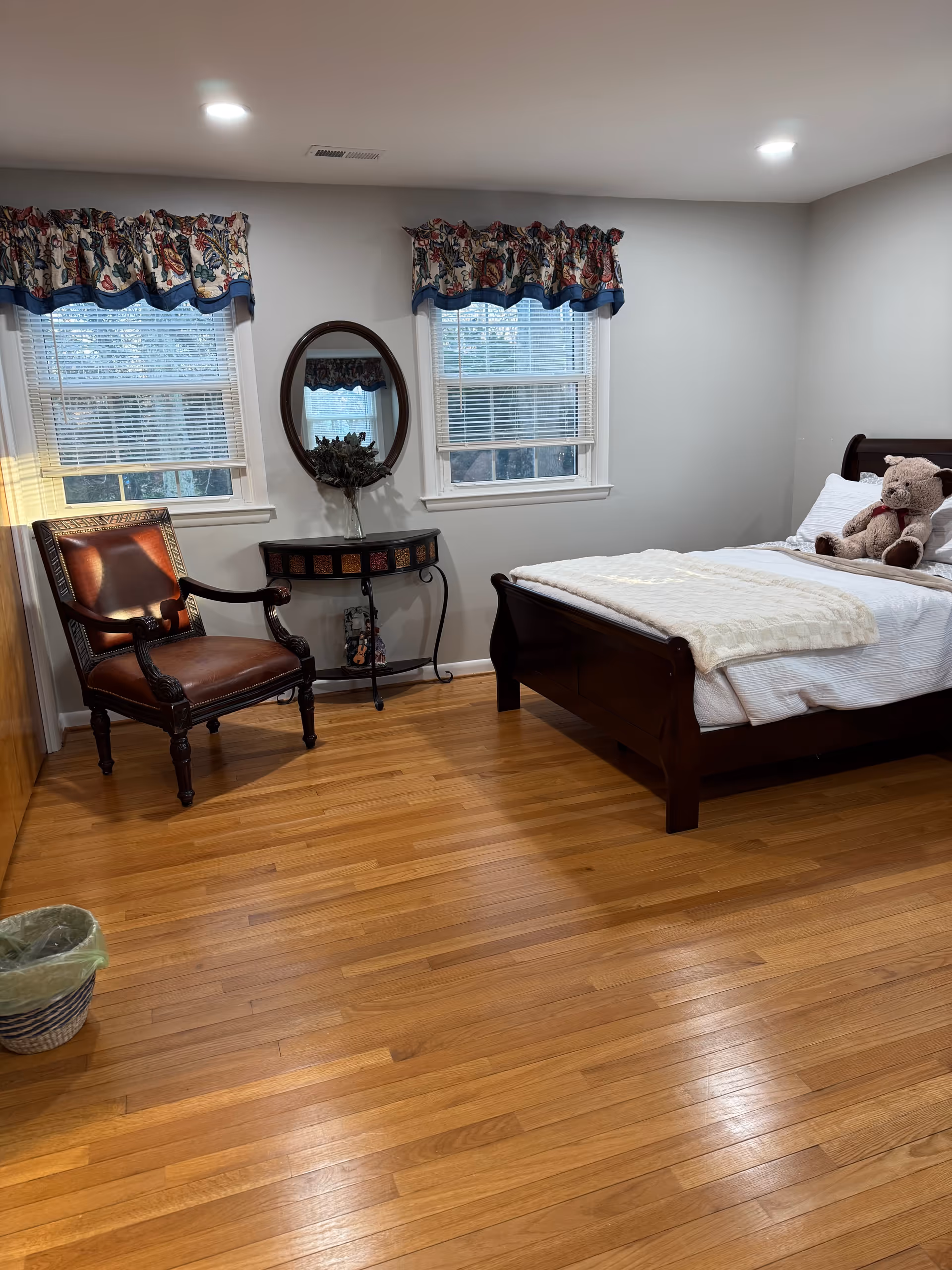 A tidy bedroom with a wooden bed, chair, round mirror console, two windows, and teddy bear.