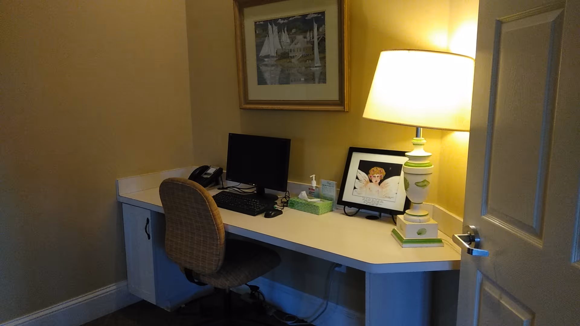 Interior home office desk with computer, lamp, framed photos, and chair against a yellow wall.