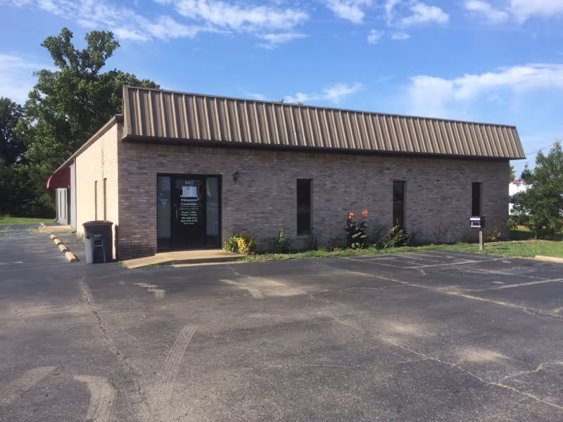 Brick building with metal roof, entrance door, and surrounding parking lot.