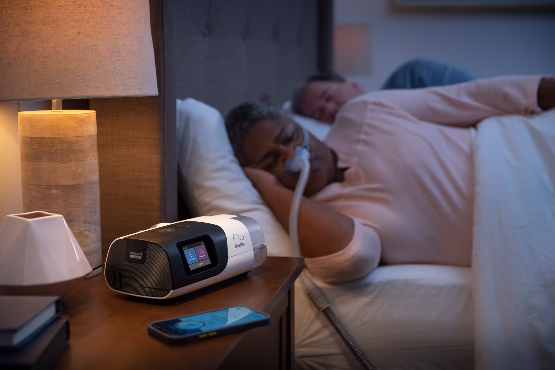 Residents rest in bed with oxygen mask; CPAP device on nightstand beside lamp.