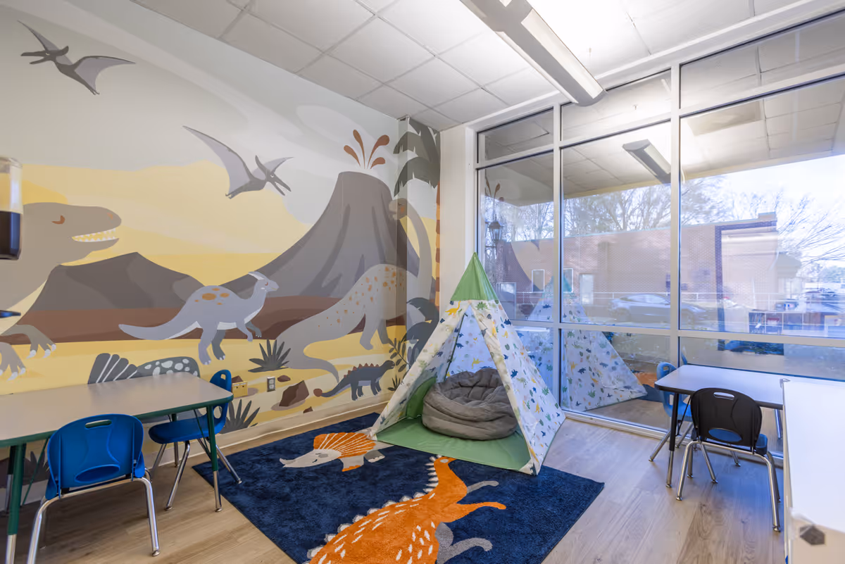 Colorful activity room with dinosaur mural, teepee tent, and small tables and chairs.