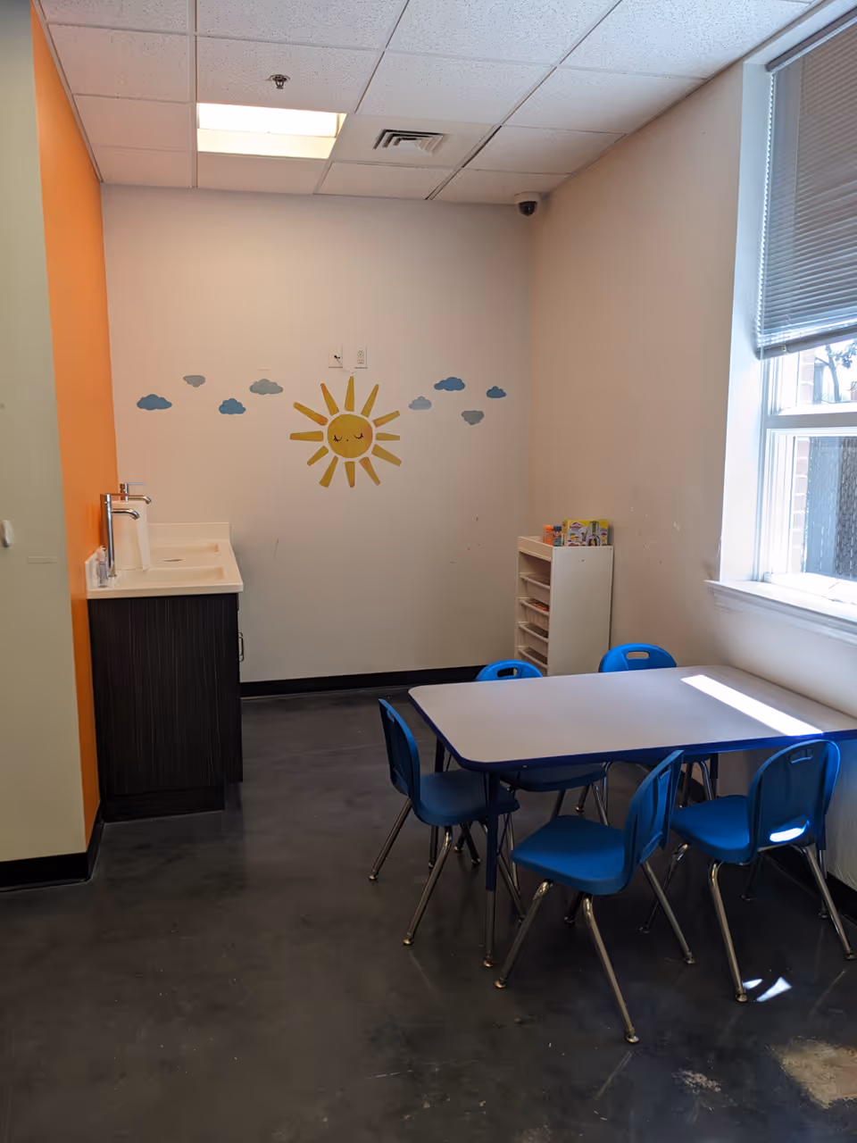 Small interior activity room with blue table and chairs, sun mural on wall.