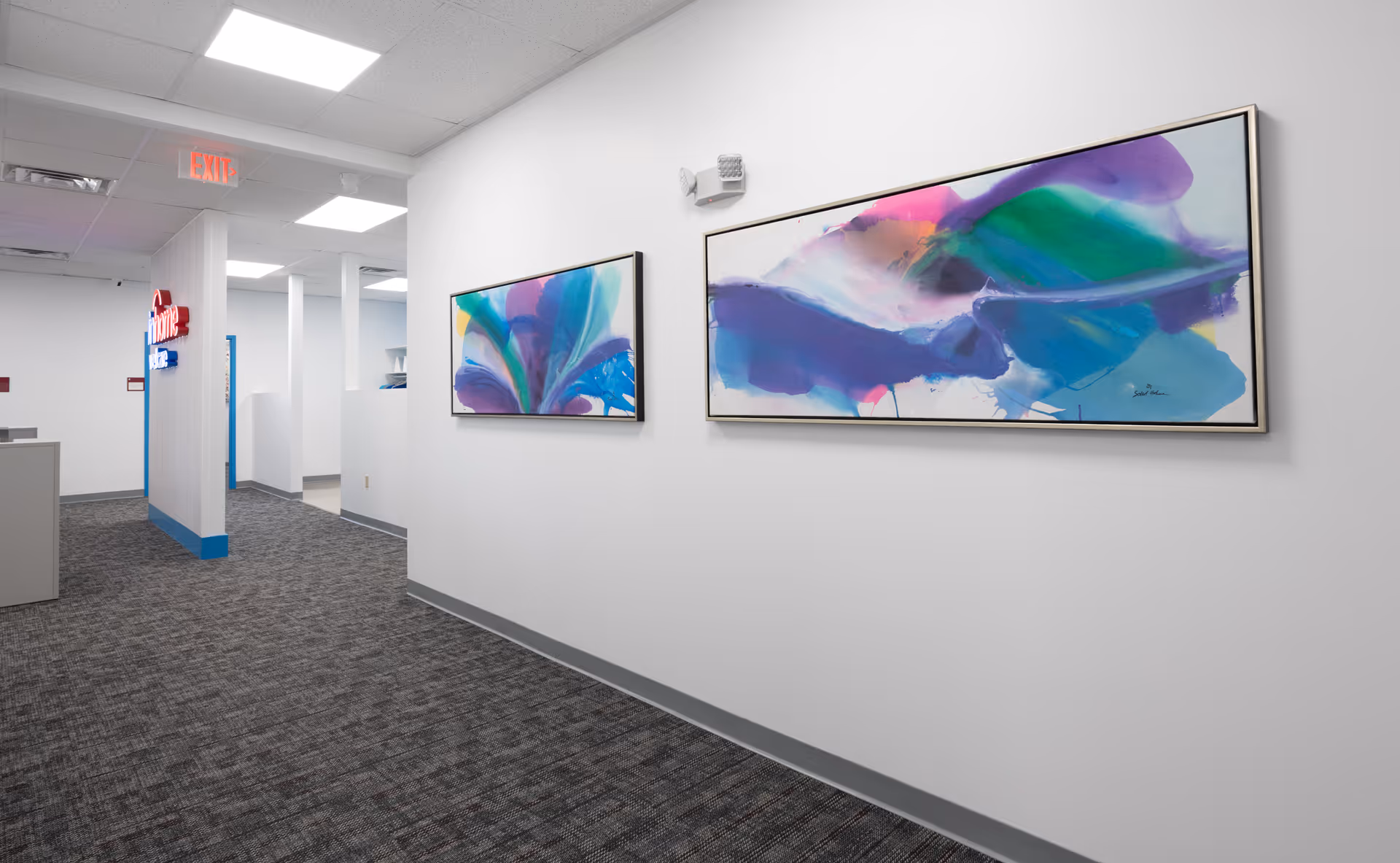 White-walled hallway in a senior facility with abstract artworks, carpeted floor, and an exit sign.