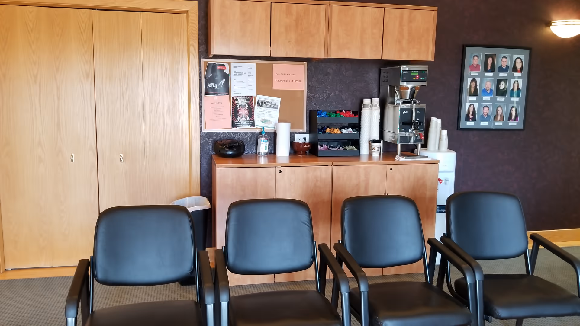 Interior break area with coffee station, cabinets, and row of black chairs.