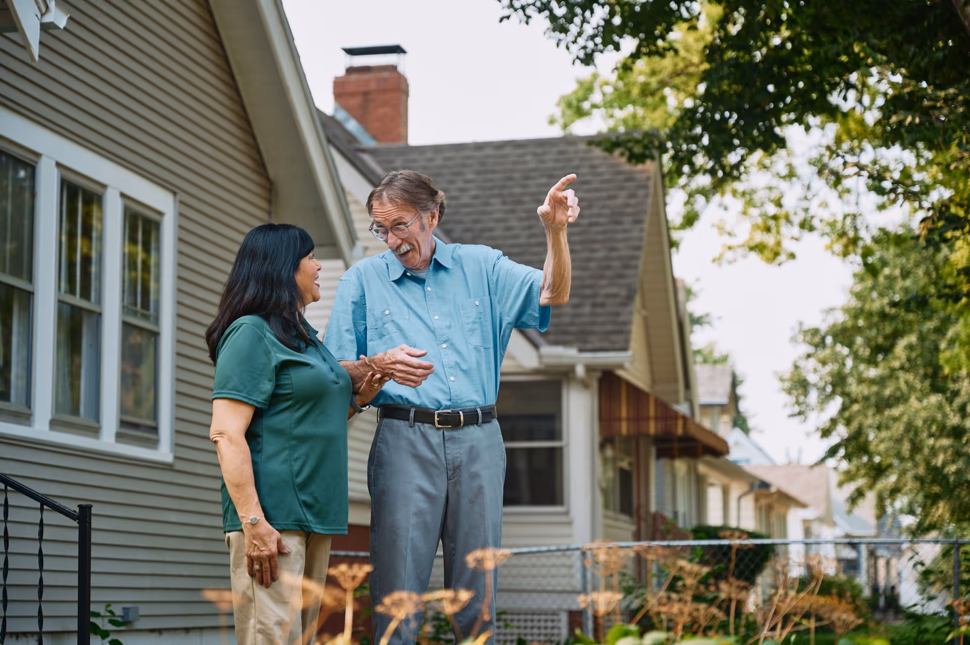 Two seniors chat outdoors beside a row of houses, smiling.