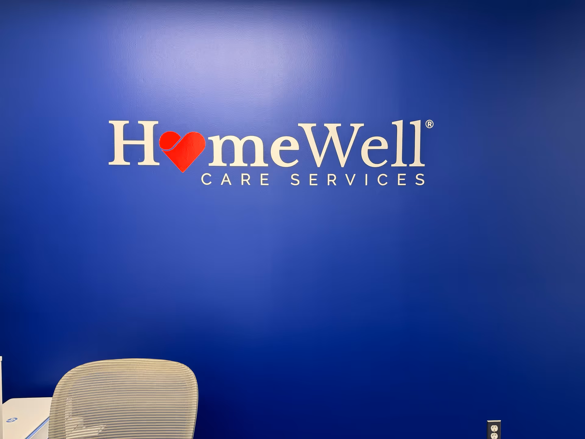 Blue interior wall with HomeWell Care Services logo and a chair in foreground.