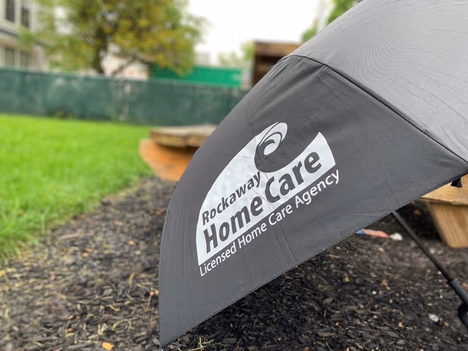 Grey umbrella with Rockaway Home Care logo in a mulched outdoor yard.