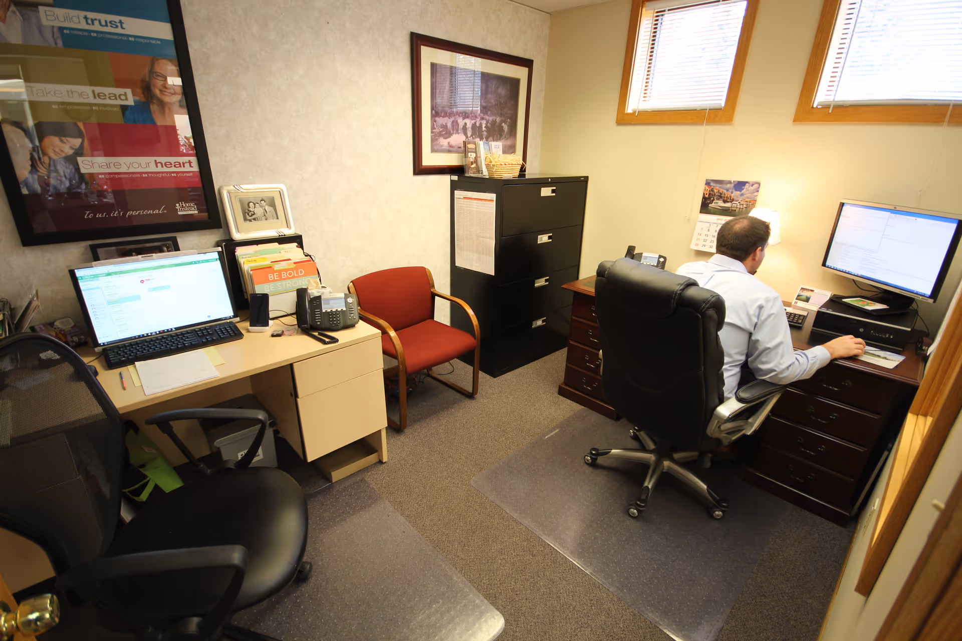 A man sits at a desk with two monitors in a small office.