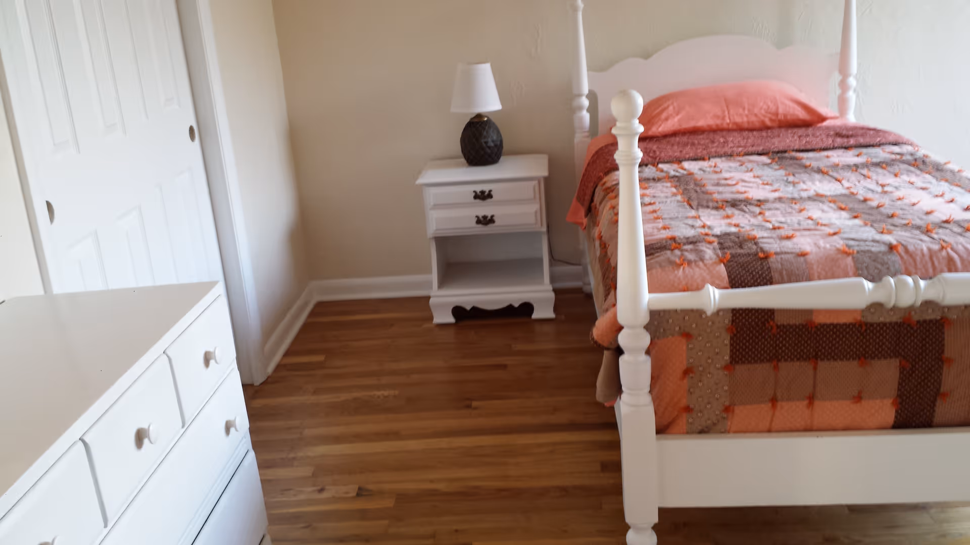 A white four-poster bed with quilt, nightstand, and dresser in a bright bedroom.