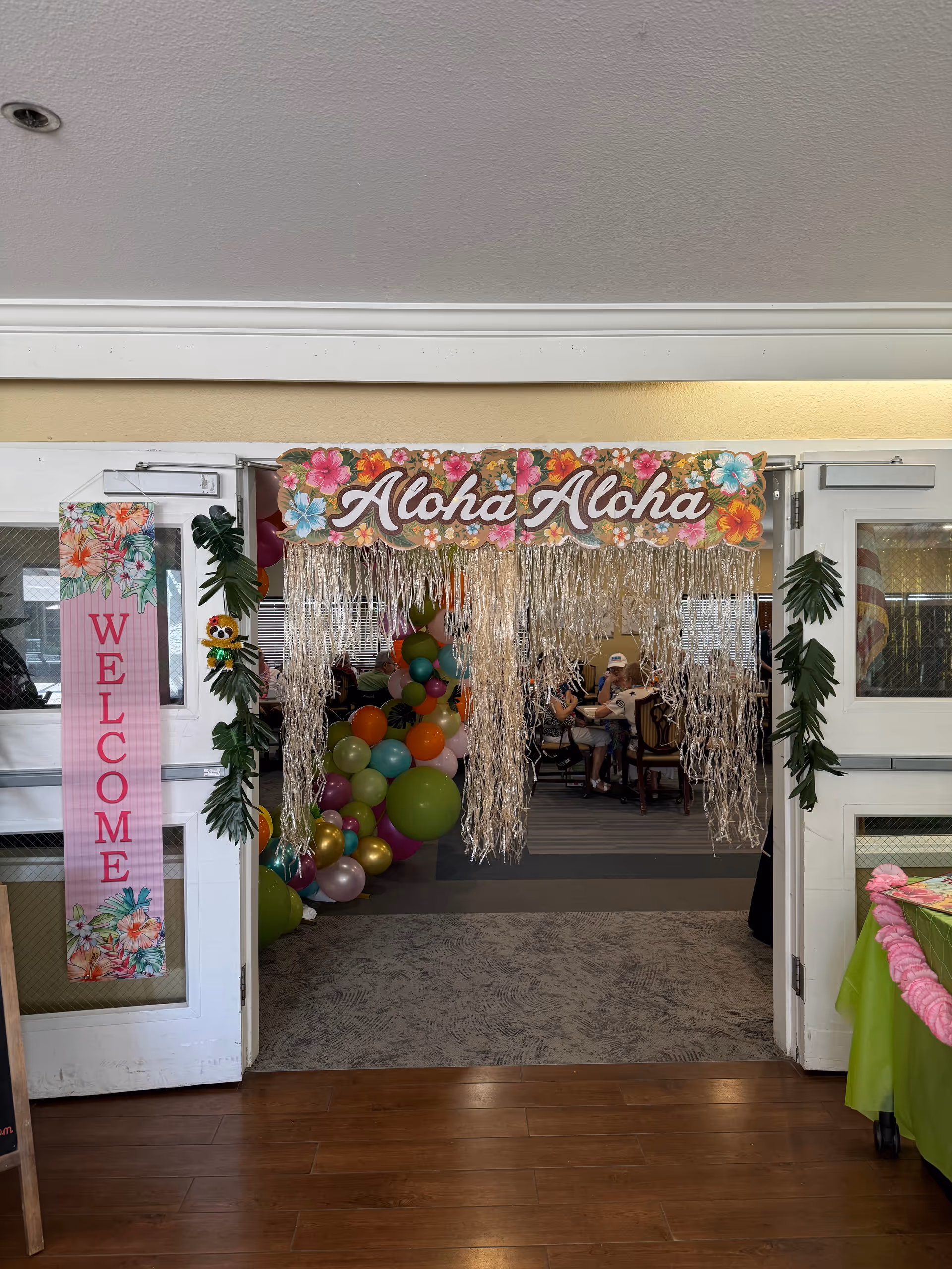 Indoor event space entrance with aloha sign, balloons, streamers, and a welcome banner.