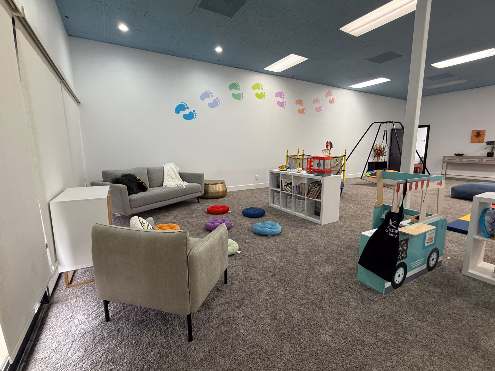 A spacious indoor play area with couches, toys, shelves, and colorful wall decals.