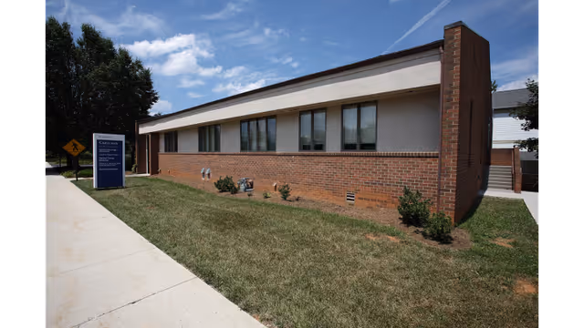 Exterior view of a low brick building with beige upper facade, windows, sidewalk, and sign.