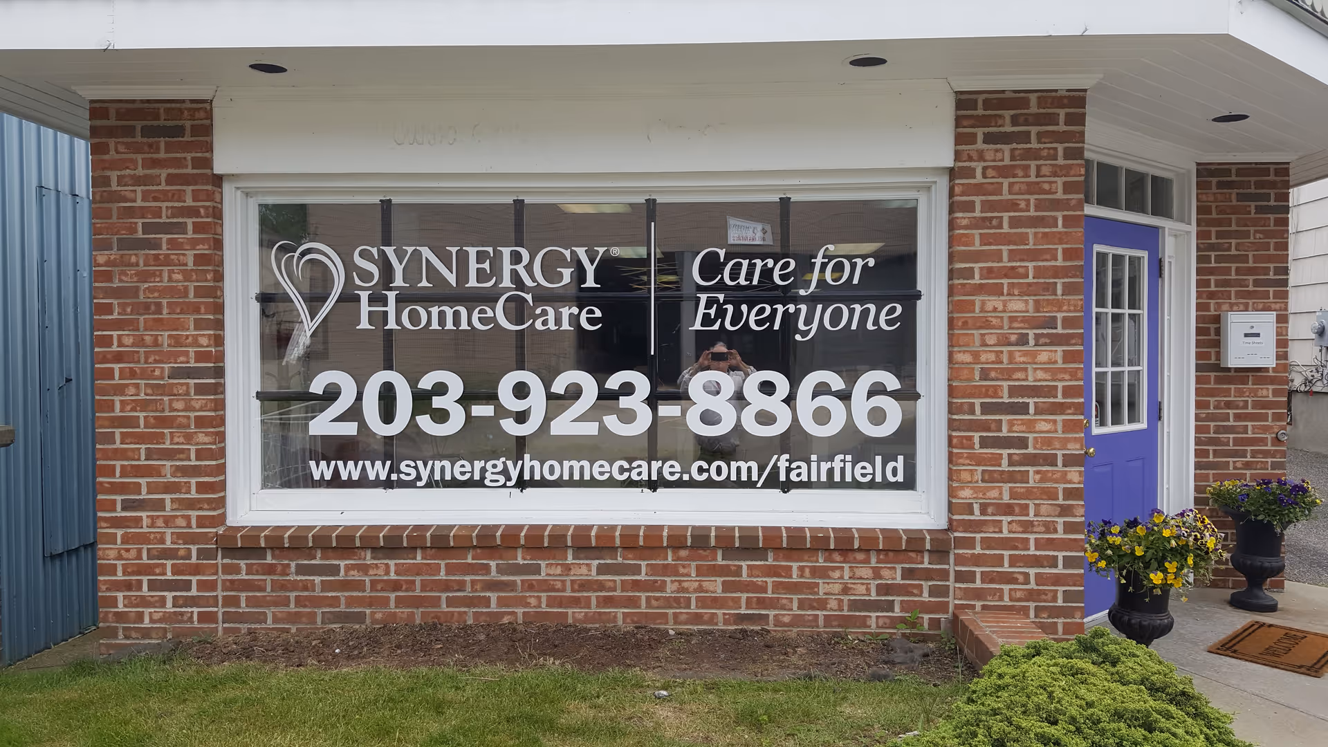 Brick storefront with Synergy HomeCare sign, large window, purple door, and planters.