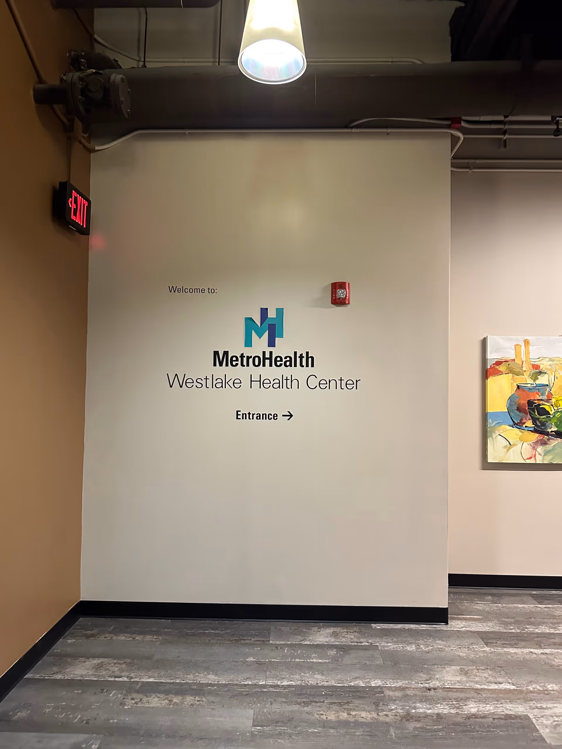Indoor corridor with MetroHealth Westlake Health Center entrance sign, exit sign, and wall artwork.
