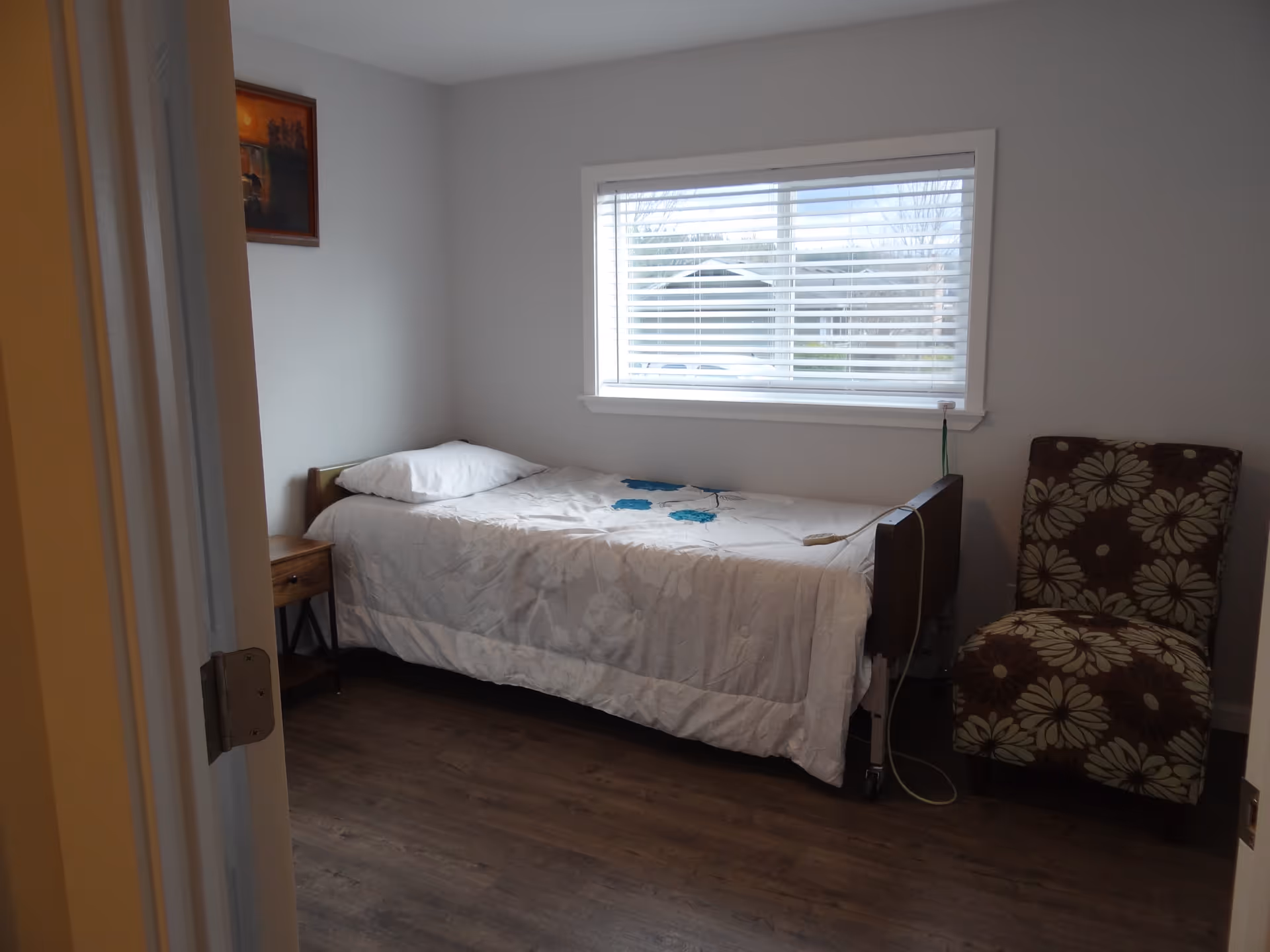 Small bedroom with a single bed, nightstand, window, and floral chair.
